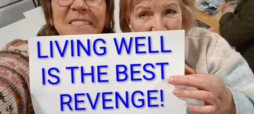 two women peeping over a sign they are holding which reads "Living well is the best revenge"